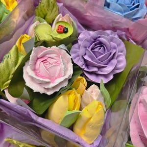 May include: Close-up of colorful floral arrangements featuring pastel-colored roses, tulips, and other flowers. The bouquets are wrapped in purple tissue paper and clear plastic. A small ladybug decoration is visible on one of the leaves.