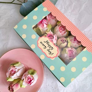 May include: A box of pink and white artificial roses with green leaves, displayed on a pink plate. The box is light blue with white polka dots and a pink scalloped edge. The box has a label that says "Happy every day!"