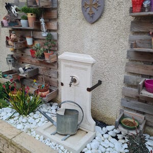 May include: A white stone fountain with a metal faucet and a watering can in front of it. The fountain is set against a wall with wooden shelves holding plants and decorative items.