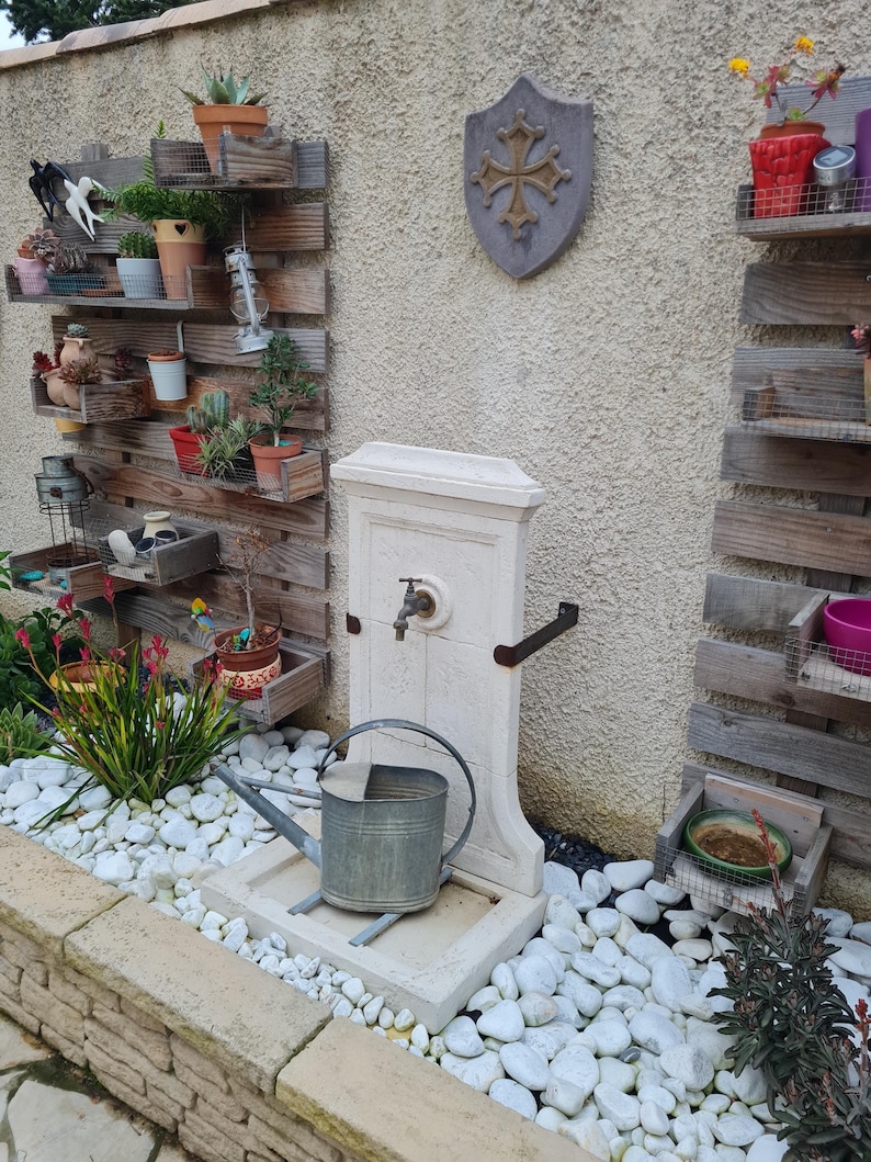 May include: A white stone fountain with a metal faucet and a watering can in front of it. The fountain is set against a wall with wooden shelves holding plants and decorative items.