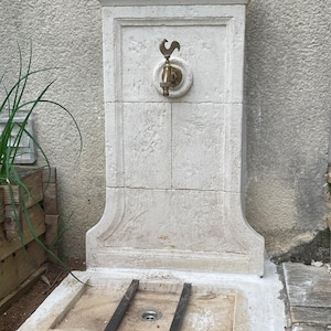 May include: A white stone outdoor water fountain with a brass faucet and a rooster design. The fountain has a square base and a rectangular basin.