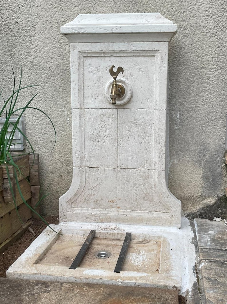 May include: A white stone outdoor water fountain with a brass faucet and a rooster design. The fountain has a square base and a rectangular basin.