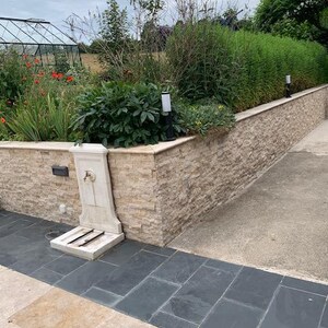 May include: A white stone fountain with a spout and two footrests sits in front of a stone wall. The wall is made of stacked stone tiles in a light beige color. The fountain is surrounded by a paved area with dark gray and light beige tiles.