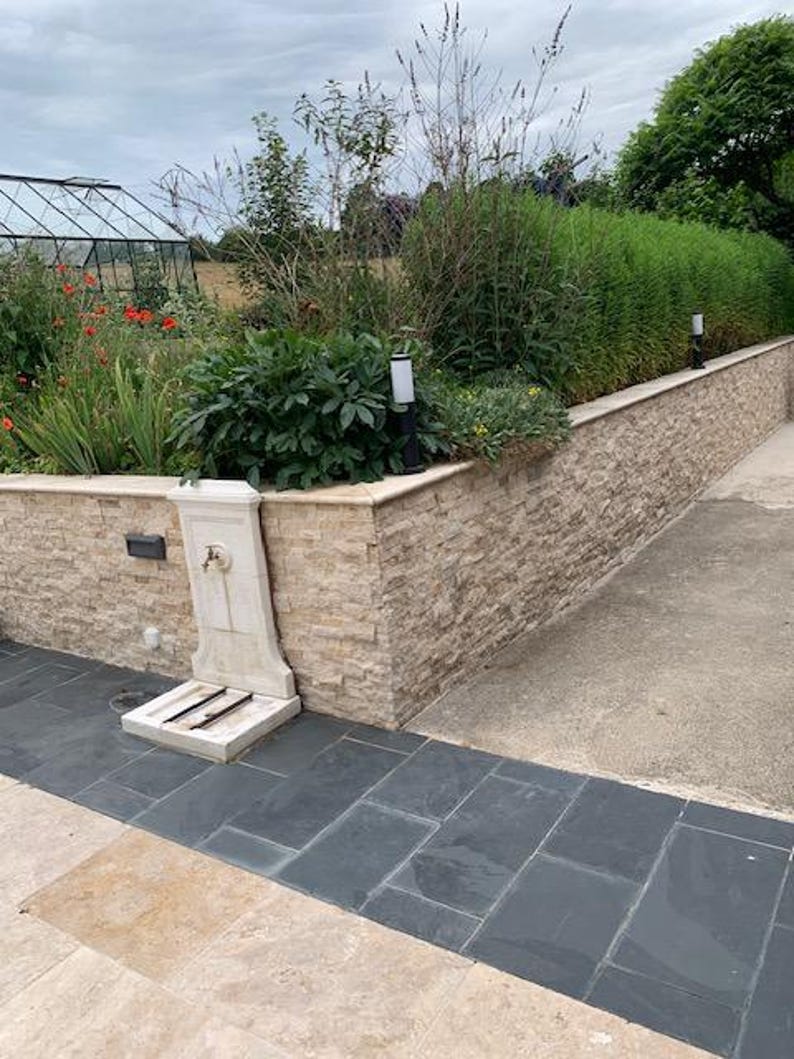 May include: A white stone fountain with a spout and two footrests sits in front of a stone wall. The wall is made of stacked stone tiles in a light beige color. The fountain is surrounded by a paved area with dark gray and light beige tiles.