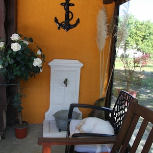 May include: A white ceramic outdoor fountain with a decorative metal anchor on the wall behind it. A potted plant with white flowers is in the foreground.