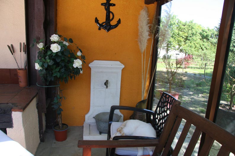 May include: A white ceramic outdoor fountain with a decorative metal anchor on the wall behind it. A potted plant with white flowers is in the foreground.