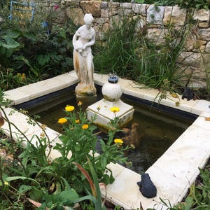 Peut inclure: Un bassin de jardin carré avec une bordure en pierre claire, comprenant une fontaine centrale blanche et une statue féminine debout tenant un récipient. Le bassin contient de petites figurines d'animaux, dont une grenouille et une tortue.