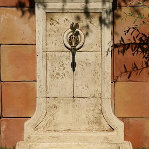 May include: A white stone wall fountain with a decorative brass faucet. The fountain has a rectangular shape with a rounded top and a small ledge at the bottom. The fountain is set against a brick wall and surrounded by green grass.