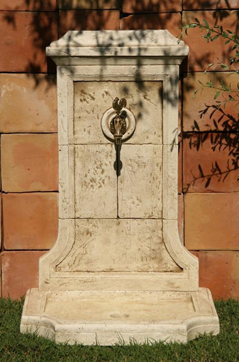 May include: A white stone wall fountain with a decorative brass faucet. The fountain has a rectangular shape with a rounded top and a small ledge at the bottom. The fountain is set against a brick wall and surrounded by green grass.