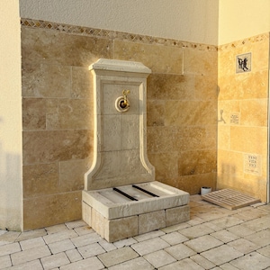 May include: A stone fountain with a gold faucet and a stone basin. The fountain is set against a wall of beige tile.
