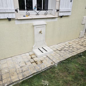 May include: A white stone outdoor water fountain with a brass faucet and a small, rectangular base. The fountain is mounted on a wall and has a decorative design.