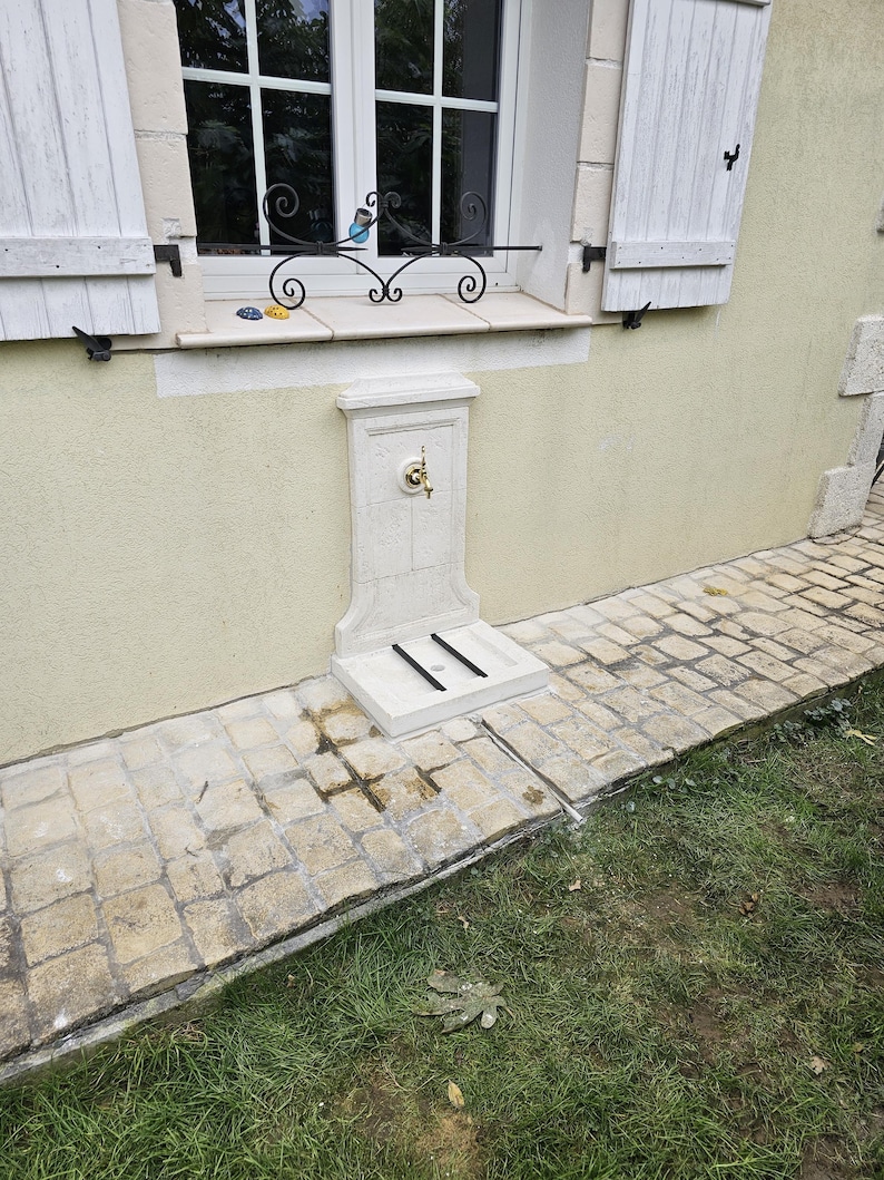 May include: A white stone outdoor water fountain with a brass faucet and a small, rectangular base. The fountain is mounted on a wall and has a decorative design.