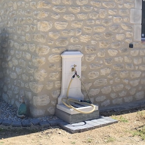 May include: A white stone outdoor water fountain with a faucet and two hoses attached. The fountain is mounted on a stone wall with a rough texture.