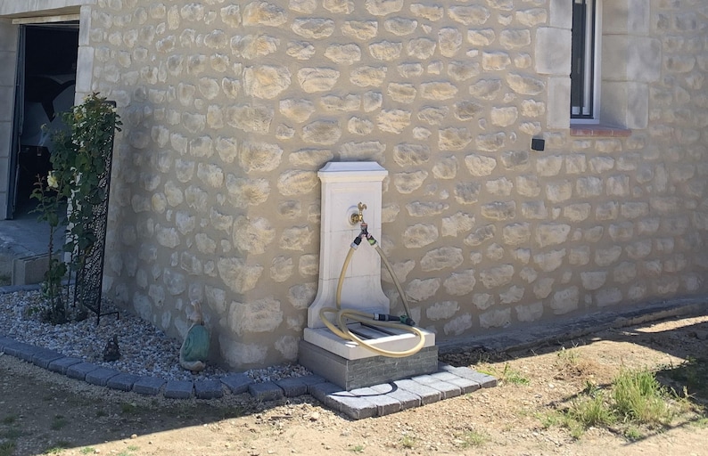 May include: A white stone outdoor water fountain with a faucet and two hoses attached. The fountain is mounted on a stone wall with a rough texture.