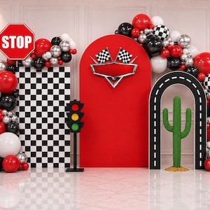 May include: A racing-themed party backdrop featuring a red arched panel with a silver emblem and two checkered flags, a black and white checkered rectangular panel, a traffic light, a stop sign, a road arch, a green cactus, and balloon garlands.