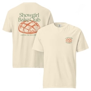 Op de afbeelding: Twee crèmekleurige t-shirts. Eén shirt heeft "Showgirl Bake Club" en een broodillustratie op de rug. Het andere shirt heeft een broodillustratie en de tekst "Loafing Him Was Bread" op de voorkant.