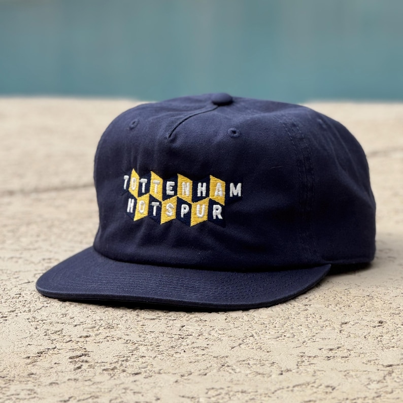May include: A navy blue baseball cap featuring an embroidered design on the front. The embroidery consists of yellow and white geometric shapes arranged in a zigzag pattern, with the text TOTTENHAM HOTSPUR stitched in white across the center.