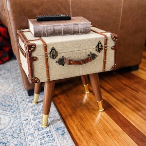 Può includere: Un tavolino laterale in stile vintage con esterno in tessuto beige, finiture in pelle marrone e dettagli in ottone. Il tavolo ha una maniglia e poggia su quattro gambe in legno con punte dorate. Un libro e un telecomando sono appoggiati sopra.