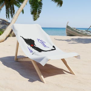 May include: A white beach chair with a towel featuring a black landmass silhouette, a red heart, and the word "Local" in blue script. The chair is on a sandy beach with a boat in the background. The scene is set by the sea.