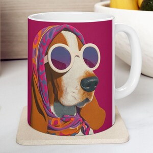 Puede incluir: Taza de cerámica blanca con un perro basset hound con gafas de sol y una bufanda colorida. El retrato del perro está sobre un fondo magenta. La taza está sobre un posavasos beige.