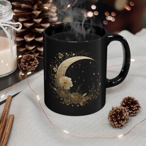 Puede incluir: Una taza de cerámica negra con un diseño de media luna con detalles florales dorados y estrellas. La taza tiene un asa curva y contiene un líquido oscuro, posiblemente té o café. Sale vapor de la taza.