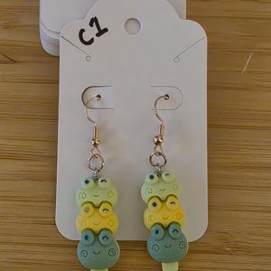 May include: A pair of whimsical frog-shaped earrings. Each earring features three stacked frog faces in shades of green, yellow, and blue, with gold-colored ear wires. The earrings are attached to a white card with the handwritten text "C1".