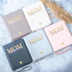 Könnte beinhalten: Fünf verschiedene farbige Reißverschlussordner mit dem Wort "MOM Organizer" in Gold auf der Vorderseite. Die Ordner sind in Schwarz, Grau, Rosa, Weiß und Braun.