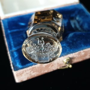 May include: A clear, faceted glass seal with a silver crest design, presented in a blue velvet box. The seal has an oval base with detailed silverwork and a gold-toned top. The crest features a shield, stars, and decorative flourishes.
