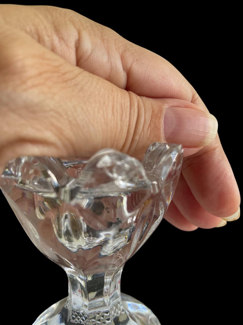 May include: A clear, cut-glass pedestal dish with a scalloped rim. The dish has a diamond pattern on the base and a faceted stem. The dish is held against a black background.