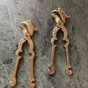 May include: A pair of ornate gold-toned nutcrackers with a bird design. The nutcrackers are detailed with intricate patterns and have a hinged jaw.