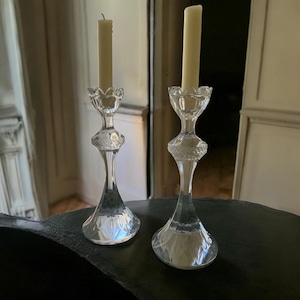May include: Two clear glass candle holders with a decorative design. Each holder has a tall, slender stem and a flared base. A cream-coloured candle sits atop each holder. The candle holders are on a dark surface.
