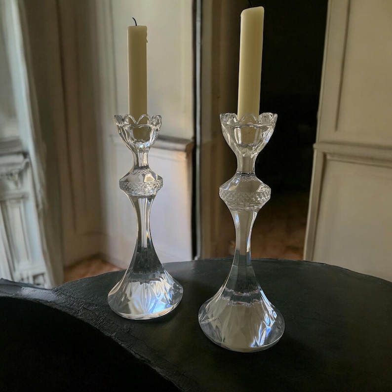 May include: Two clear glass candle holders with a decorative design. Each holder has a tall, slender stem and a flared base. A cream-coloured candle sits atop each holder. The candle holders are on a dark surface.