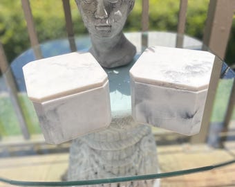 May include: Two white marble-patterned hexagonal boxes with lids sit on a glass table. A gray statue of a woman's head with flowers in her hair is behind the boxes. The boxes have a gray and white veined pattern.