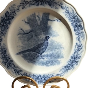 May include: A white ceramic plate with a blue pheasant and tree scene. The plate has a scalloped edge with a blue floral and acorn design. The plate is displayed on a gold-colored metal stand.