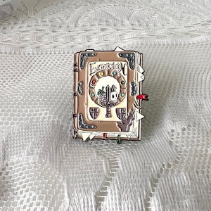 May include: A brown book-shaped enamel pin with a skull and sword design. The book has a white cover with the text "Enchiridion" in gold lettering. The pin has a red, white, and blue color scheme.