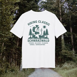 Könnte beinhalten: Weißes T-Shirt mit grünem Aufdruck eines Wanderers, Bäumen und Sonne, mit dem Text "HIKING CLASSIC SCHWARZWALD" und Ortsnamen. Ein T-Shirt zum Thema Natur, ideal für Wanderfreunde.