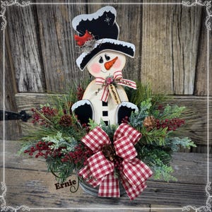 May include: A wooden snowman decoration with a black top hat and a red cardinal. The snowman has a carrot nose and a red and white striped scarf. It sits in a metal container filled with greenery, red berries, and a red and white checkered bow.