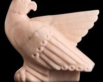 May include: A pale pink stone sculpture of an eagle in profile, featuring a curved beak, a single visible wing extended outward, and carved circular details along the primary feathers, set against a solid black background.