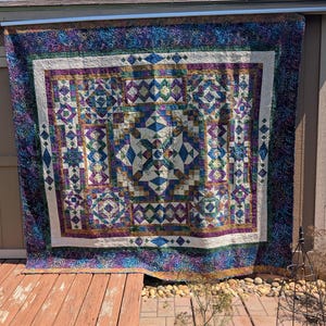 May include: A large, colorful quilt with a geometric pattern, displayed outdoors. The quilt features a central design of diamonds and triangles in shades of blue, purple, and gold, framed by a patterned border. The quilt is hanging on a wall.