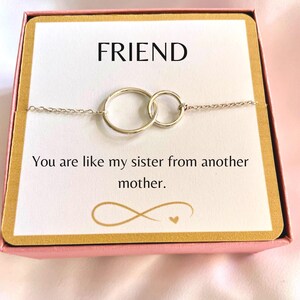 May include: A silver friendship bracelet with two interlocking circles, presented in a gift box with a card that reads "FRIEND You are like my sister from another mother."