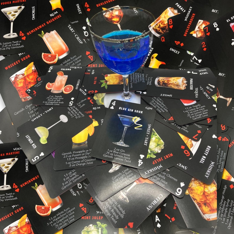 May include: A close-up of a pile of playing cards with cocktail recipes. The cards are black with red and white text. The recipes include drinks like the Hemingway Daiquiri, Whiskey Sour, Blue Gin Sour, Mint Julep, and Rusty Nail. The cards are arranged in a random order, with some overlapping.