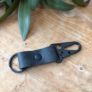 Puede incluir: Un llavero de cuero negro con un mosquetón de metal negro y un llavero redondo. La correa de cuero se asegura con un broche negro. El llavero está sobre una superficie de madera.
