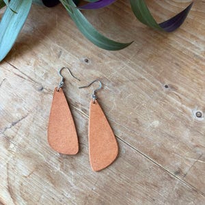 Puede incluir: Un par de pendientes de cuero naranja con ganchos plateados. Los pendientes tienen forma de lágrima con una superficie texturizada. Se exhiben sobre una superficie de madera, con follaje verde y morado en el fondo.