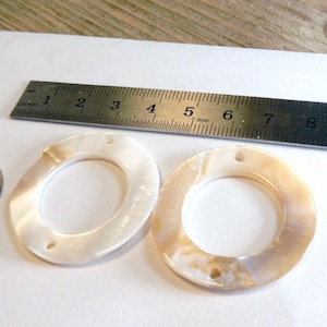 May include: Two oval-shaped, iridescent, mother-of-pearl beads with two holes each. A ruler shows measurements in millimeters. A coin is next to the beads. The beads are on a white surface.