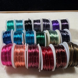 May include: A collection of spools of colorful craft wire in various metallic shades. The spools are arranged in rows, showcasing a spectrum of colors including green, blue, purple, pink, and bronze. The wire is tightly wound on white plastic spools.