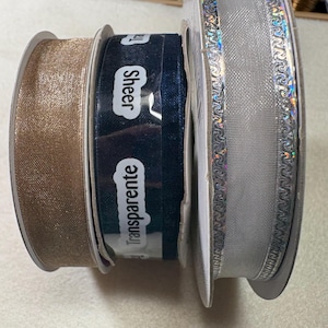 May include: Spools of decorative ribbon in various colors. Includes a gold, sheer navy blue, and silver ribbon. The navy blue ribbon has the word "Sheer" and "Transparente" printed on it.