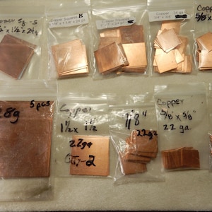 May include: Several small plastic bags filled with copper squares of various sizes and gauges. Handwritten labels indicate dimensions in inches and weight in grams. Some bags are labeled "Copper Squares" and "Copper". The copper pieces have a reddish-brown color.