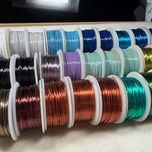May include: A collection of spools of colorful craft wire in various shades, including silver, blue, purple, green, and copper. The wire is tightly wound on white spools, creating a visually appealing display. Ideal for jewelry making and crafting.