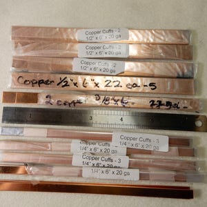 May include: An overhead shot shows several copper strips, some labeled "Copper Cuffs," in various sizes. A metal ruler is also present. The strips are packaged and ready for use. The image is well-lit, highlighting the copper's color.