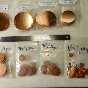 May include: An assortment of copper discs in various sizes, packaged in clear plastic bags. The sizes range from 7/16 inch to 2 inches in diameter. A metal ruler is placed between the bags for scale. The bags are labeled with the disc size and gauge.